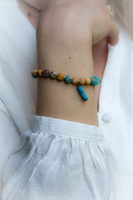 Turkusowa Oaza Bransoletka Z Jaspisu I Turkusu Ozdobna: Bransoletka z koralików z turkusu, jaspisu i złotych przekładek, z wiszącym, turkusowym walcem. Zdjęcie na tle białej, lekko rozmazanej tkaniny.