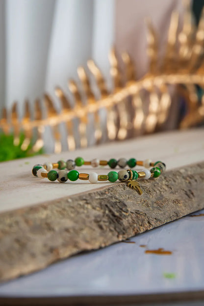 TWOJA OAZA - BRANSOLETKA Z ZIELONEGO JASPISU CESARSKIEGO I HOWLITU. Bransoletka z zielonymi i białymi koralikami, ułożona na kamieniu, tło z złotymi liśćmi.