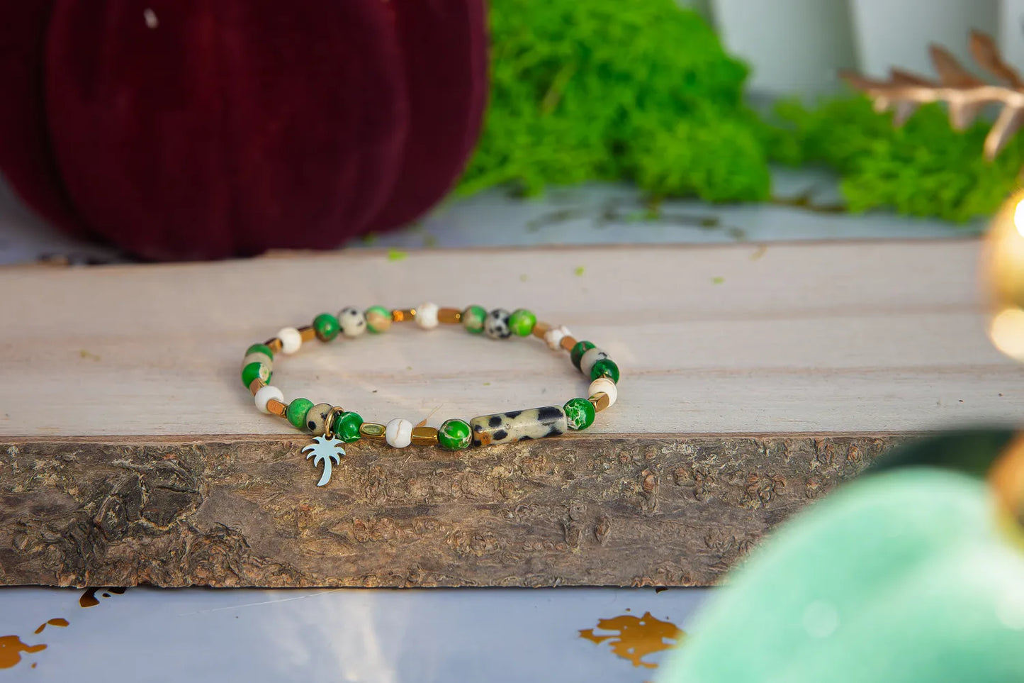 TWOJA OAZA - BRANSOLETKA Z ZIELONEGO JASPISU CESARSKIEGO I HOWLITU. Bransoletka z zielonymi i białymi koralikami, ozdobiona uroczym zawieszkiem w kształcie palmowca. Tło z drewnianym pniem, zielonym mechem i czerwonymi elementami.