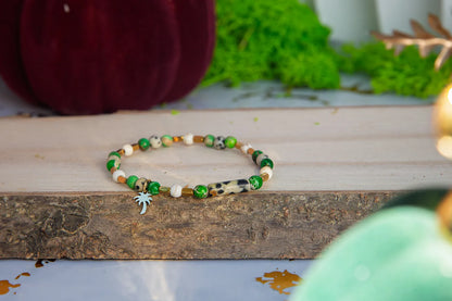TWOJA OAZA - BRANSOLETKA Z ZIELONEGO JASPISU CESARSKIEGO I HOWLITU. Bransoletka z zielonymi i białymi koralikami, ozdobiona uroczym zawieszkiem w kształcie palmowca. Tło z drewnianym pniem, zielonym mechem i czerwonymi elementami.