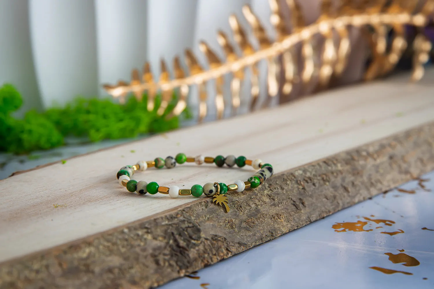 TWOJA OAZA - BRANSOLETKA Z ZIELONEGO JASPISU CESARSKIEGO I HOWLITU. Bransoletka z kolorowymi koralikami zielonego jaspisu cesarskiego i howlitu, ułożona na drewnianym tle z zielonymi roślinami i złotym liściem w tle.