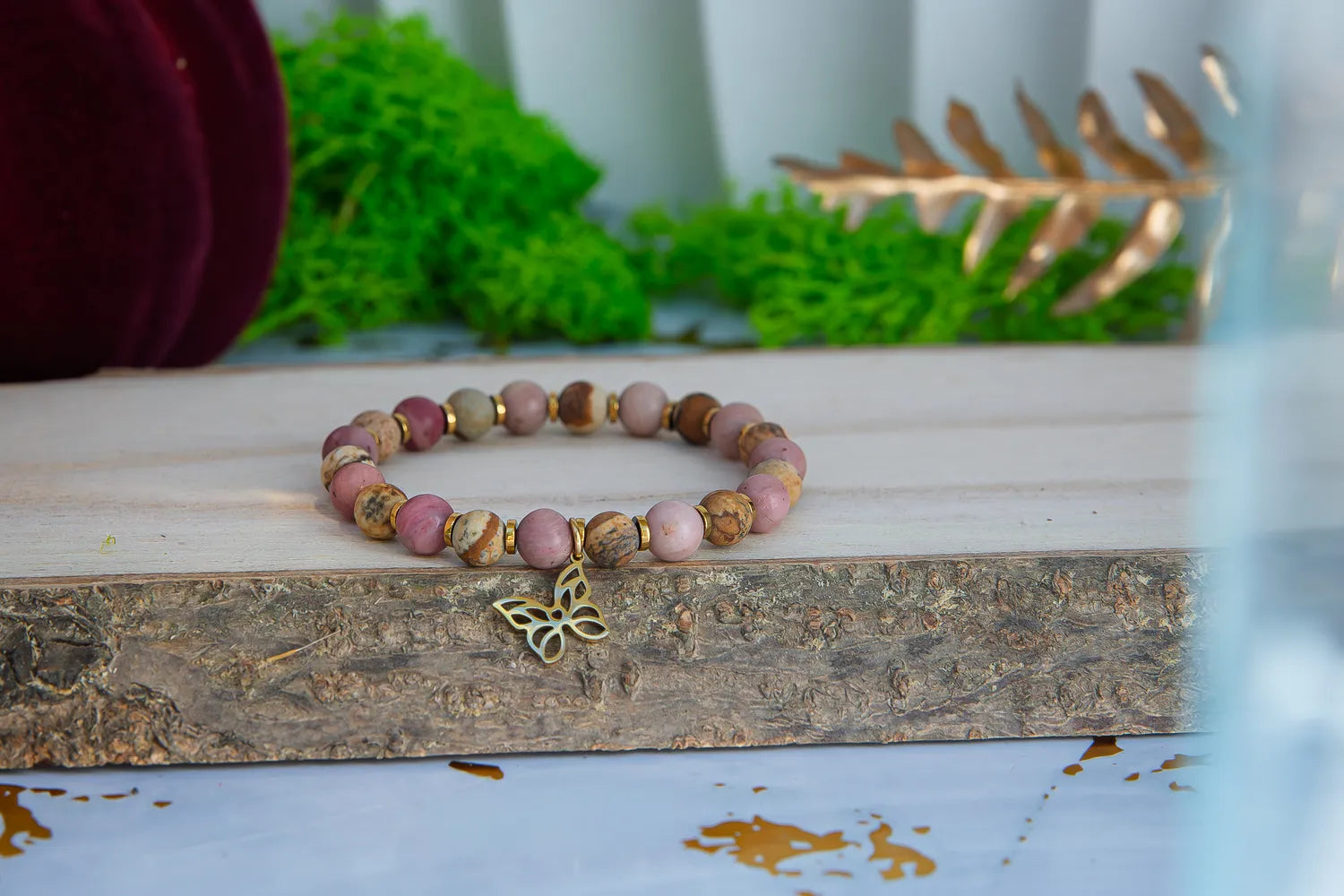 ULECZONE SKRZYDŁA - BRANSOLETKA Z RODONITU I JASPISU Z MOTYLEM. Bransoletka z kolorowymi koralikami z rodonitu i jaspisu, ozdobiona złotym motylem, leży na drewnianym tle z zielonymi roślinami w tle.