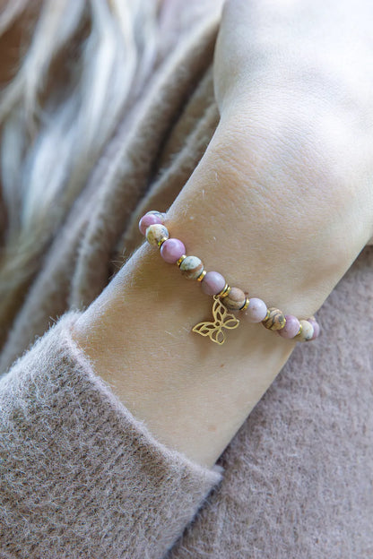 ULECZONE SKRZYDŁA - BRANSOLETKA Z RODONITU I JASPISU Z MOTYLEM. Bransoletka z kolorowymi koralikami z rodonitu i jaspisu oraz złotym motylem na tle delikatnej, beżowej tkaniny.