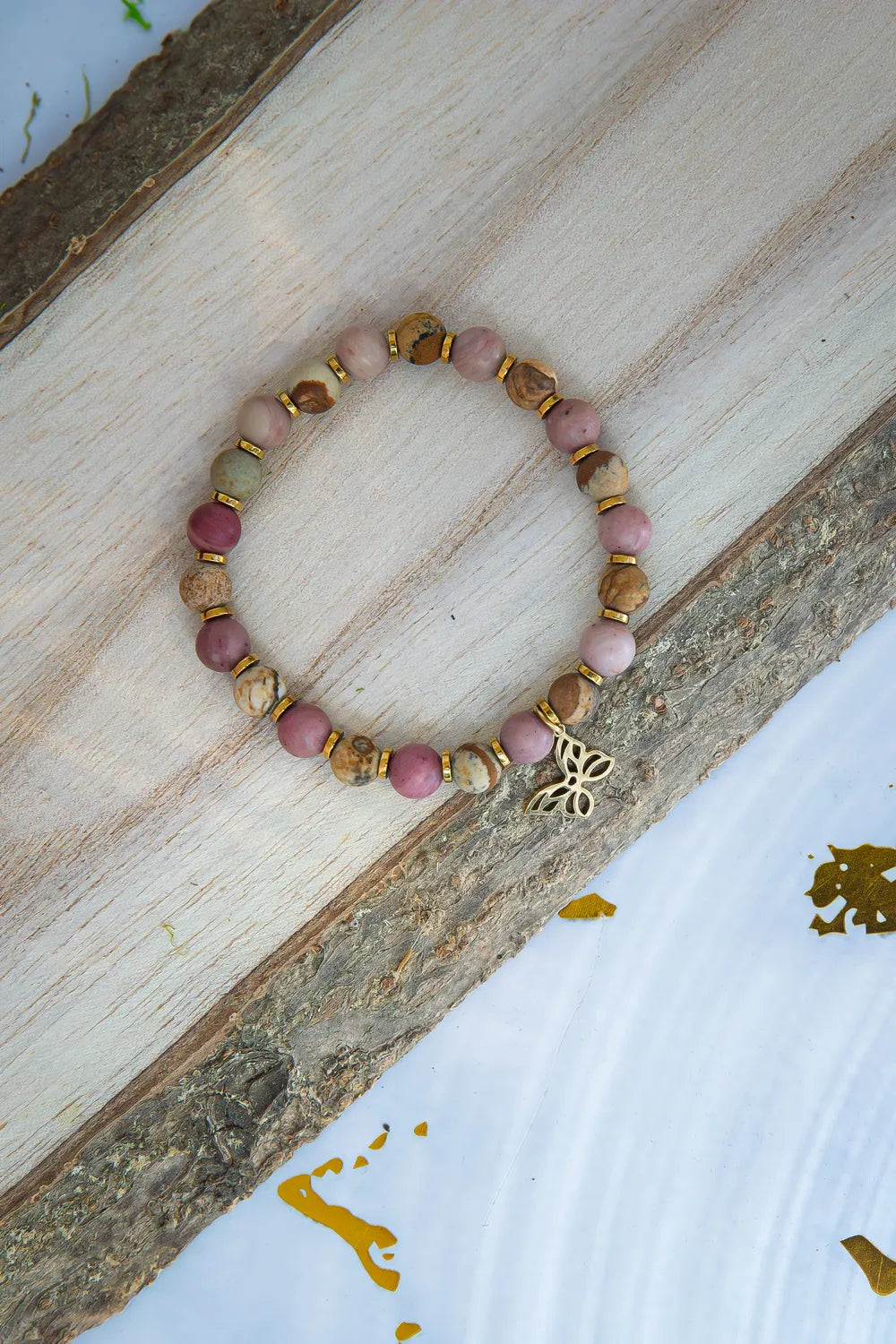 ULECZONE SKRZYDŁA - BRANSOLETKA Z RODONITU I JASPISU Z MOTYLEM. Bransoletka z różowymi i brązowymi koralikami, ozdobiona motylem, leży na drewnianym tle.
