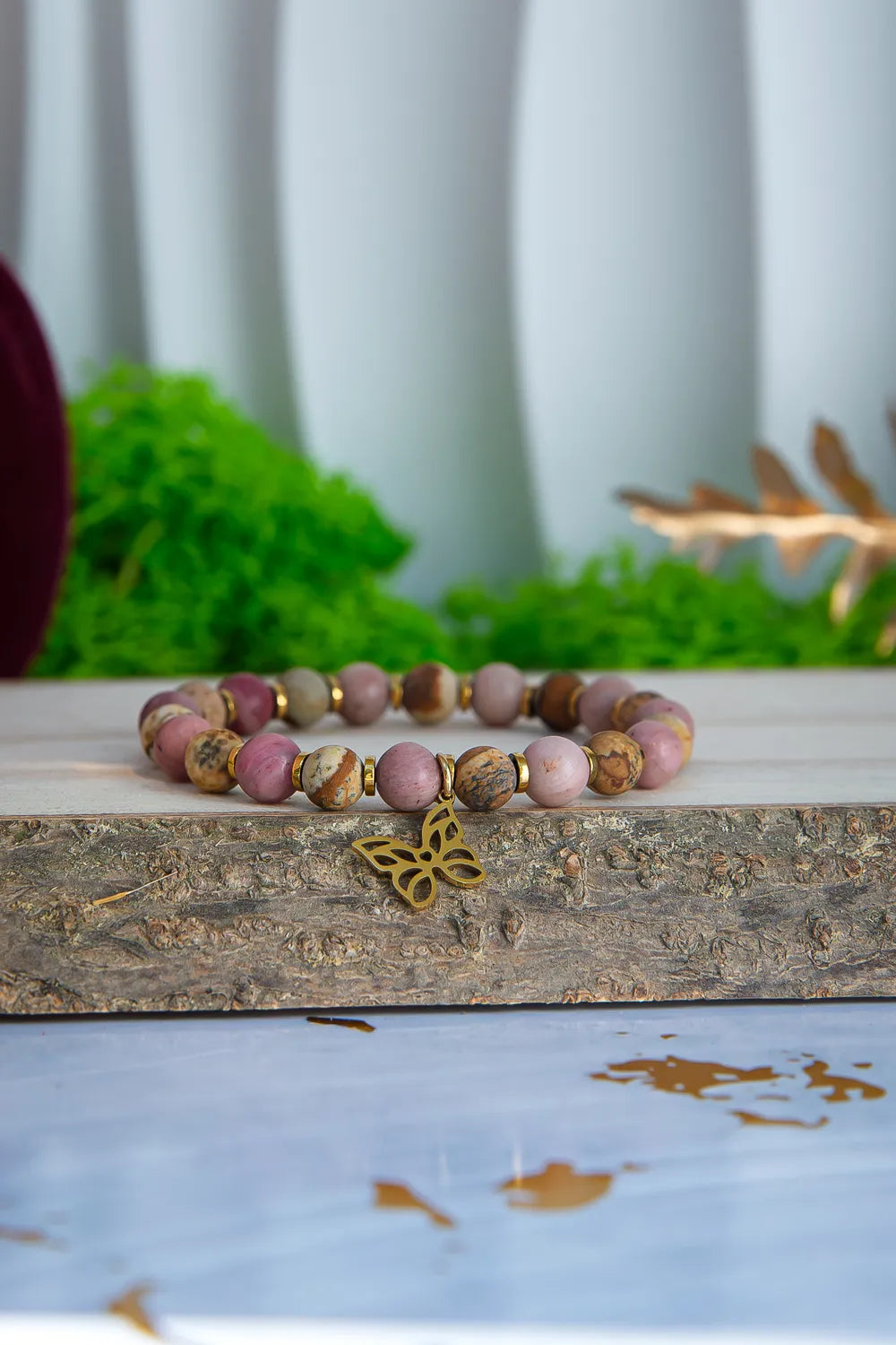 ULECZONE SKRZYDŁA - BRANSOLETKA Z RODONITU I JASPISU Z MOTYLEM. Bransoletka z koralikami z różowego i beżowego kamienia, ozdobiona złotym motylem, leży na drewnianym bloku z zielonym tłem.