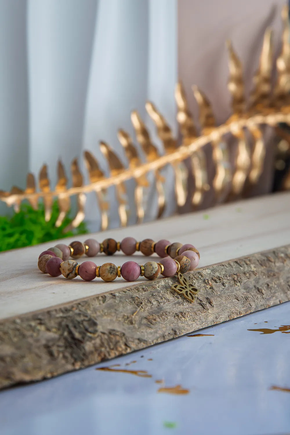 ULECZONE SKRZYDŁA - BRANSOLETKA Z RODONITU I JASPISU Z MOTYLEM. Bransoletka z koralikami z różowego rodonitu i jaspisu, ozdobiona złotym motylem, leży na drewnianym pniu z tłem z zielonymi liśćmi i złotą gałęzią.