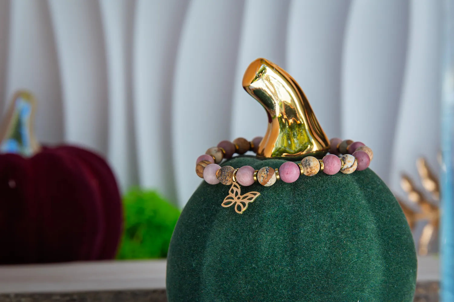 ULECZONE SKRZYDŁA - BRANSOLETKA Z RODONITU I JASPISU Z MOTYLEM. Bransoletka z różowymi i beżowymi koralikami oraz złotym motylem, umieszczona na zielonym podstawce z złotym liściem w tle.