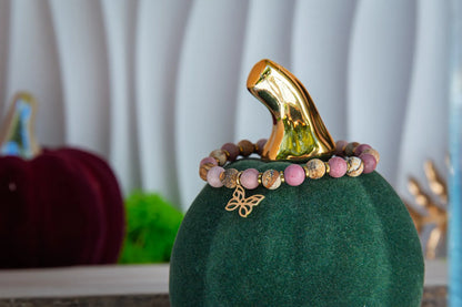 ULECZONE SKRZYDŁA - BRANSOLETKA Z RODONITU I JASPISU Z MOTYLEM. Bransoletka z różowymi i beżowymi koralikami oraz złotym motylem, umieszczona na zielonym podstawce z złotym liściem w tle.