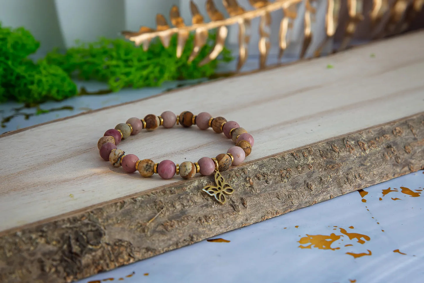 ULECZONE SKRZYDŁA - BRANSOLETKA Z RODONITU I JASPISU Z MOTYLEM. Bransoletka z koralikami z różowego rodonitu i jaspisu, ozdobiona złotym motylem, leży na drewnianym tle z zielonymi roślinami w tle.