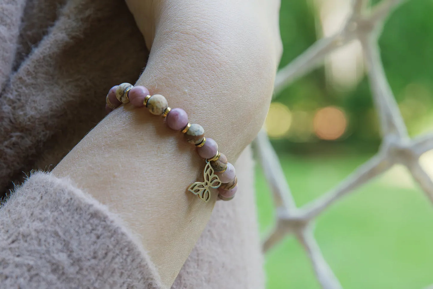 ULECZONE SKRZYDŁA - BRANSOLETKA Z RODONITU I JASPISU Z MOTYLEM. Bransoletka z kolorowymi koralikami z rodonitu i jaspisu, ozdobiona złotym motylem. Tło z zielonymi liśćmi i siatką.