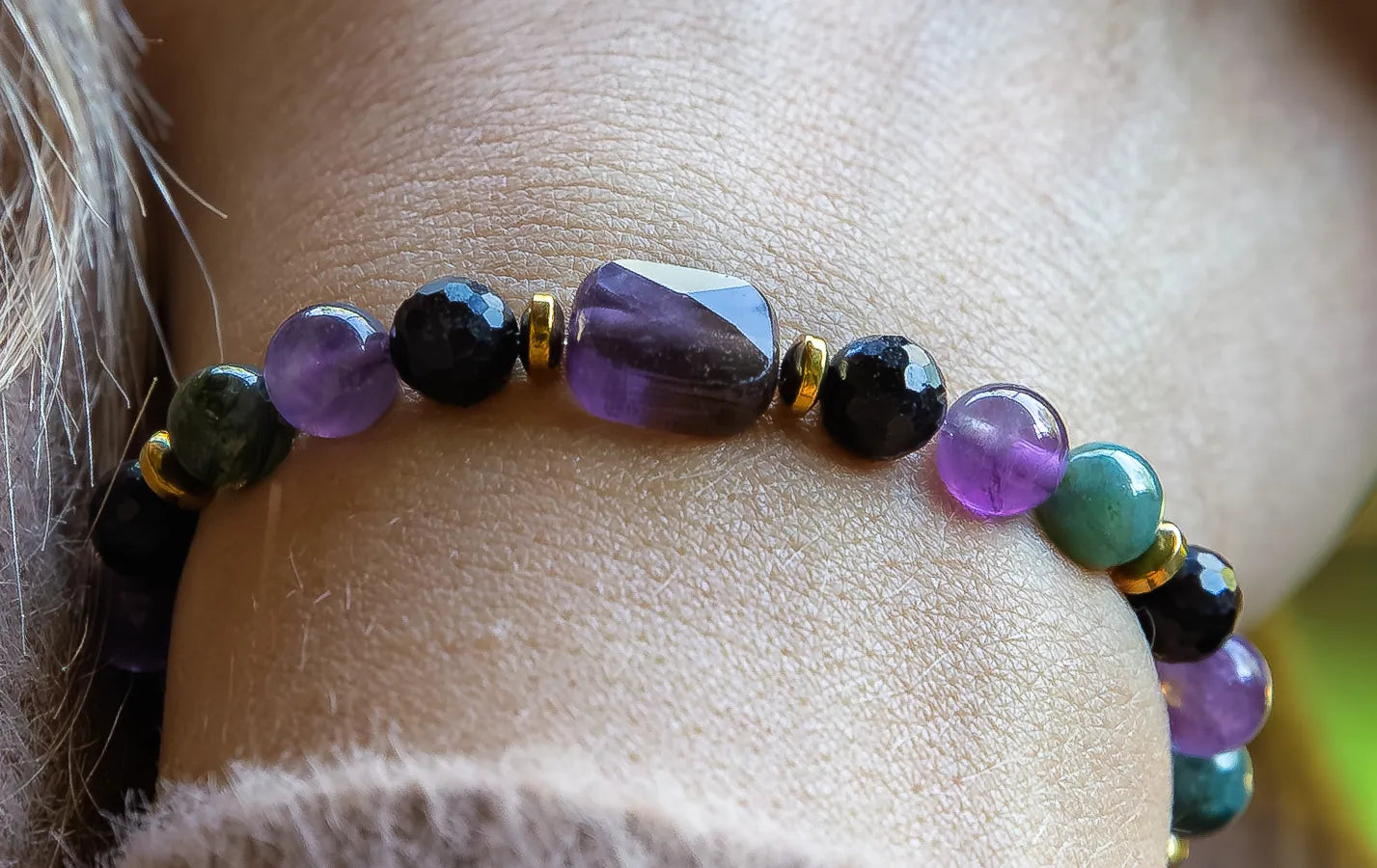 WRZOSOWA NOC - BRANSOLETKA Z AMETYSTU I AGATU INDYJSKIEGO. Bransoletka z kolorowymi kamieniami, w tym ametystem i agatem indyjskim, na tle skóry. Kamienie są ułożone w równomiernym rzędzie.