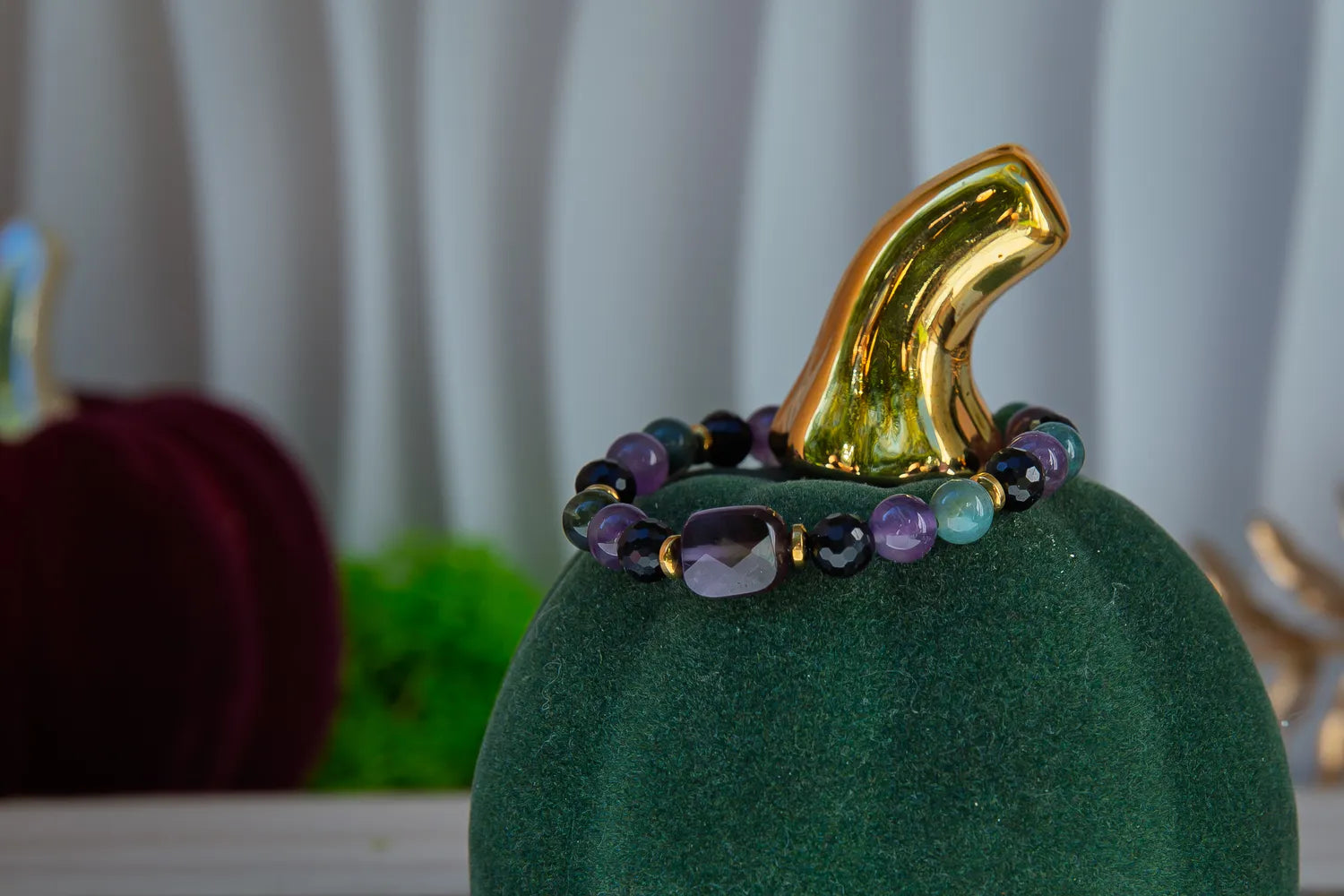 WRZOSOWA NOC - BRANSOLETKA Z AMETYSTU I AGATU INDYJSKIEGO. Bransoletka z kolorowymi kamieniami ametystu i agatu indyjskiego, ułożona na zielonym podstawce w kształcie dyni z złotym kapeluszem, na tle białych zasłon.