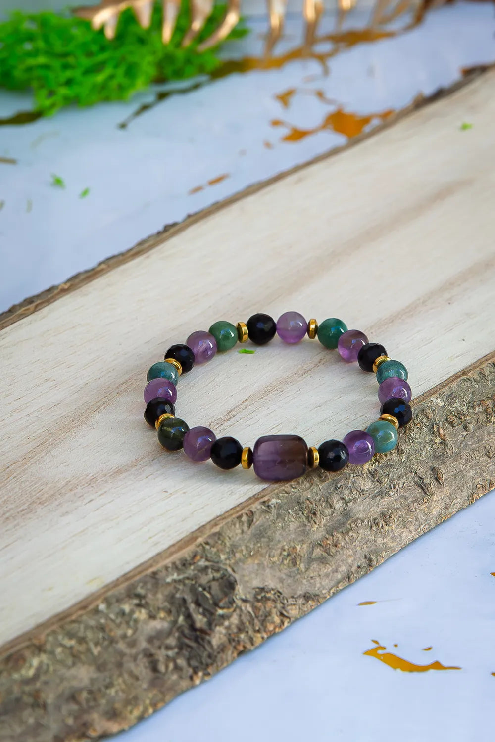 WRZOSOWA NOC - BRANSOLETKA Z AMETYSTU I AGATU INDYJSKIEGO, złożona z kolorowych kamieni, leżąca na drewnianym tle z zielonymi liśćmi w tle.