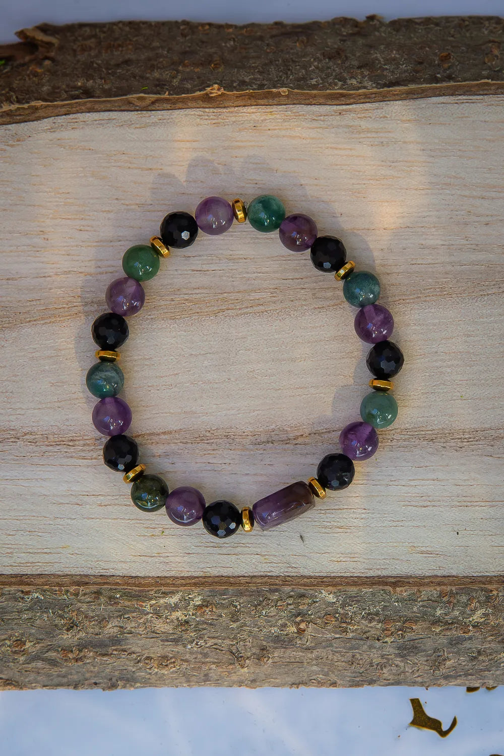 WRZOSOWA NOC - BRANSOLETKA Z AMETYSTU I AGATU INDYJSKIEGO, prezentująca się na drewnianym tle. Biżuteria składa się z kolorowych kamieni, w tym ametystu i agatu indyjskiego, ułożonych w elegancki wzór.