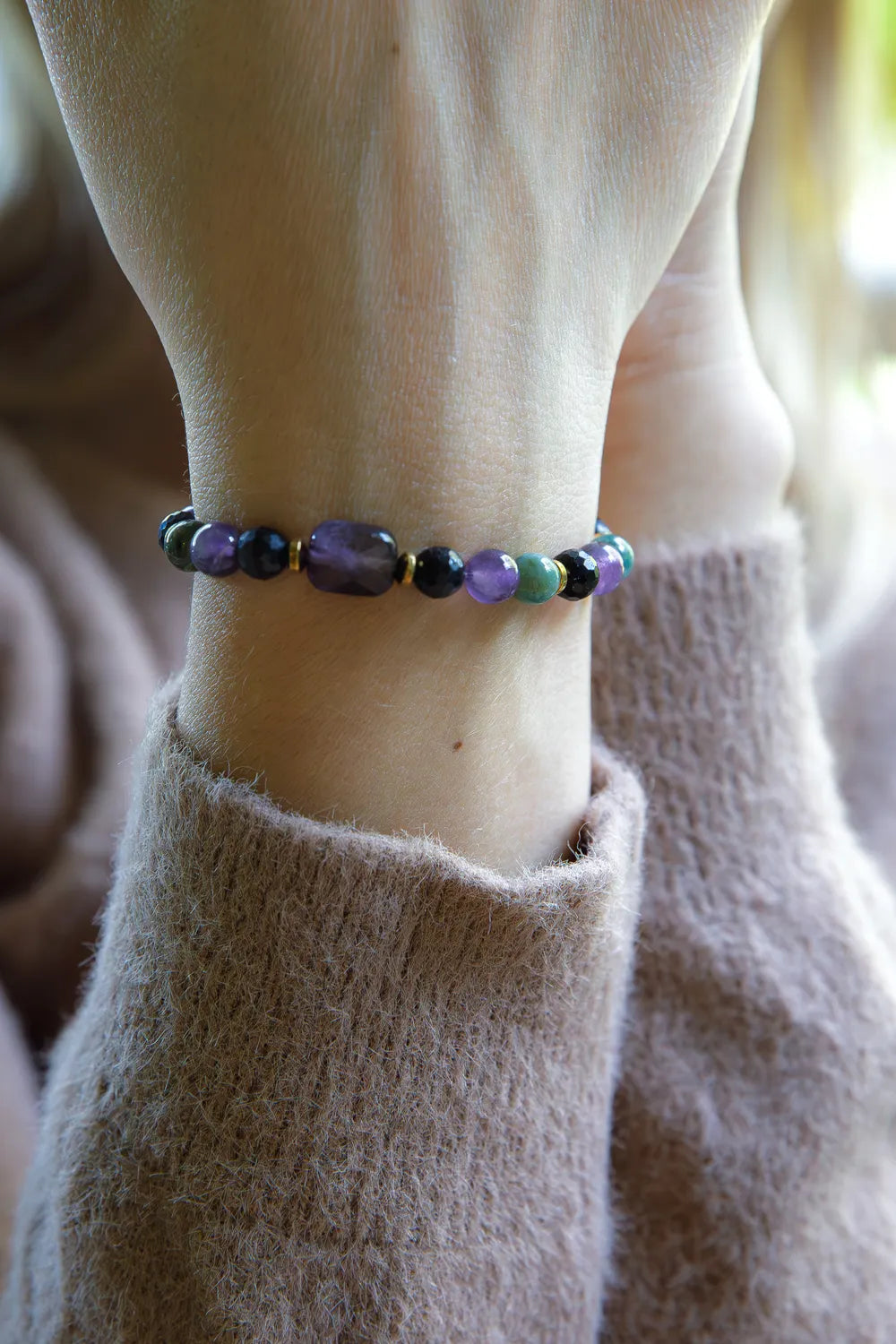 WRZOSOWA NOC - BRANSOLETKA Z AMETYSTU I AGATU INDYJSKIEGO na nadgarstku, z widocznymi kolorowymi kamieniami w odcieniach fioletu i zieleni, na tle delikatnej, beżowej tkaniny.