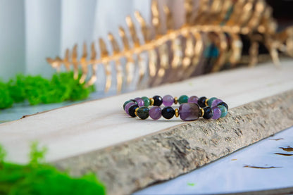 WRZOSOWA NOC - BRANSOLETKA Z AMETYSTU I AGATU INDYJSKIEGO, prezentująca się na drewnianym tle z zielonymi roślinami w tle. Biżuteria składa się z kolorowych kamieni, w tym ametystu i agatu indyjskiego.