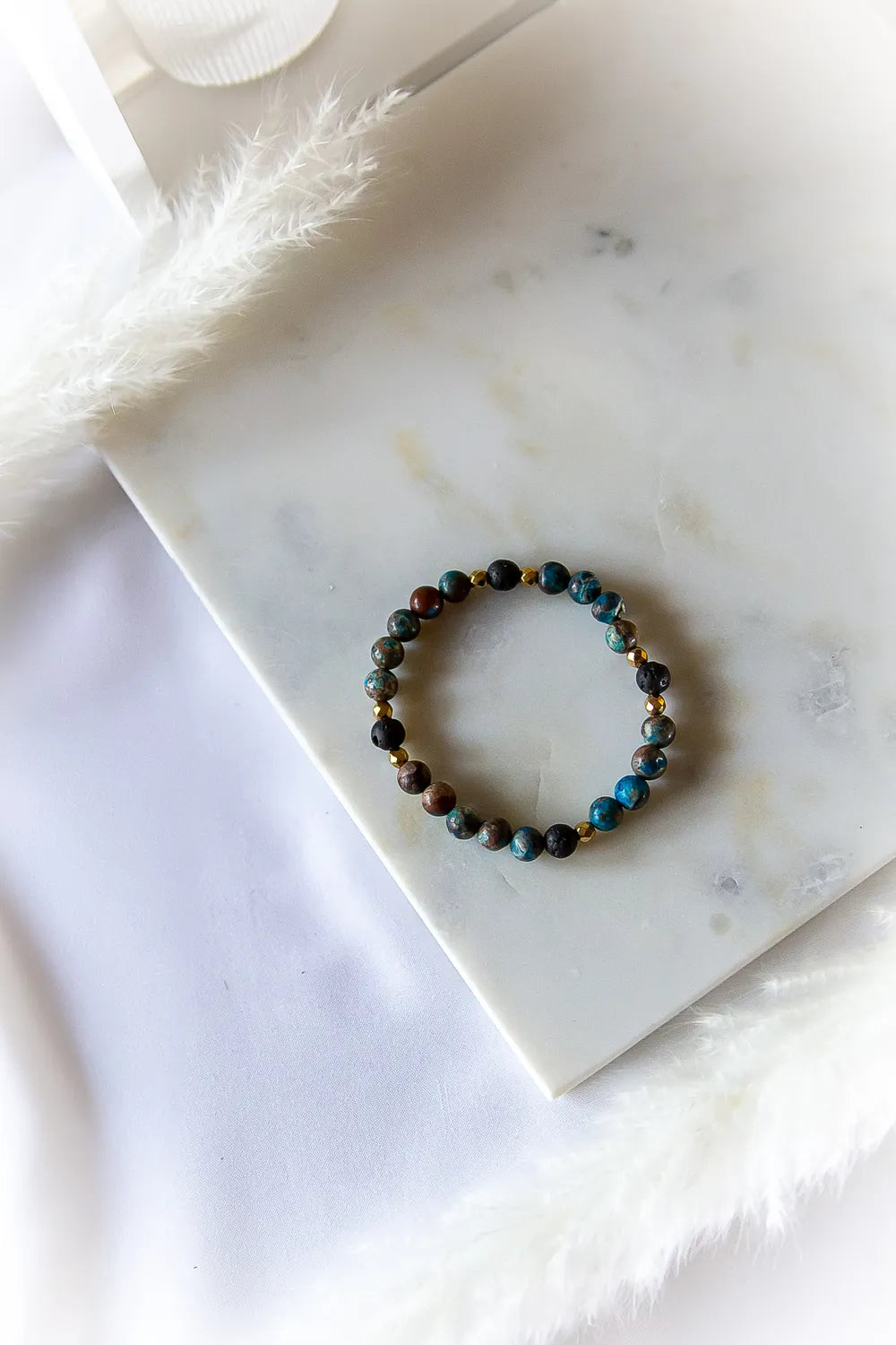 Wulkaniczna Moc Bransoletka Z Turkusem I Lawą. Bransoletka z turkusowymi, brązowymi i czarnymi kamieniami, przeplatana złotymi i czarnymi elementami, leży na białej marmurowej płycie z piórami.