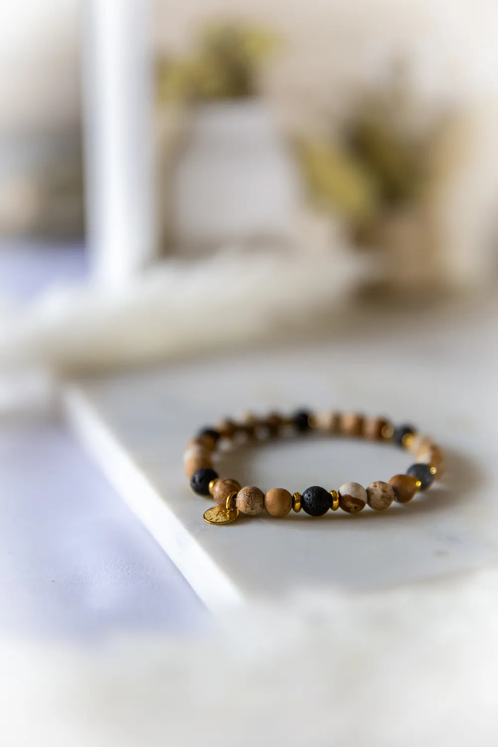 Ziemski Rytm Bransoletka Jaspis Czarna Lawa z ozdobnym, złotym zawieszeniem na białym, kamiennym tle.