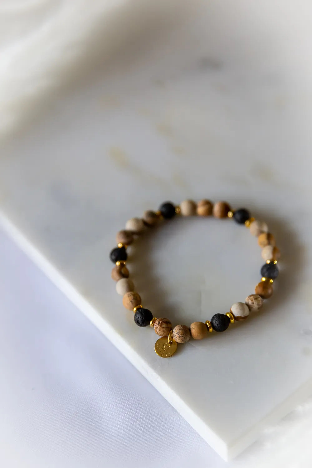 Ziemski Rytm Bransoletka Jaspis Lawa z kamieniami w odcieniach beżu, brązu i czerni, na marmurowym tle.