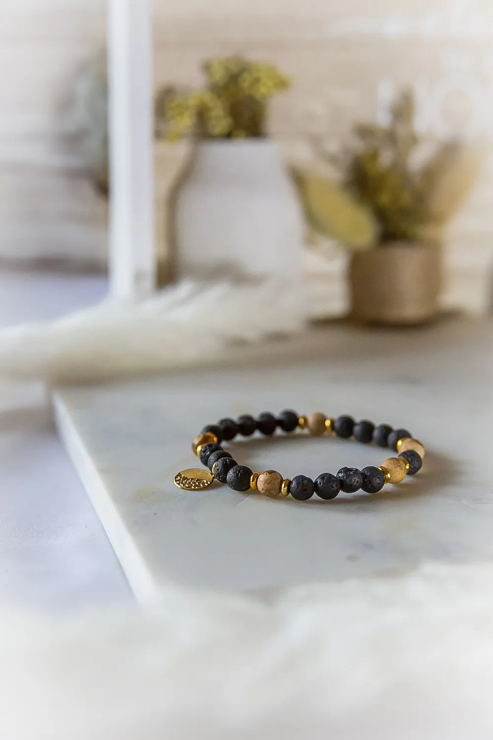 Ziemskie Ścieżki Bransoletka Jaspis Czarna Lawa na marmurowej powierzchni z złotym medalionem, w tle suche kwiaty.