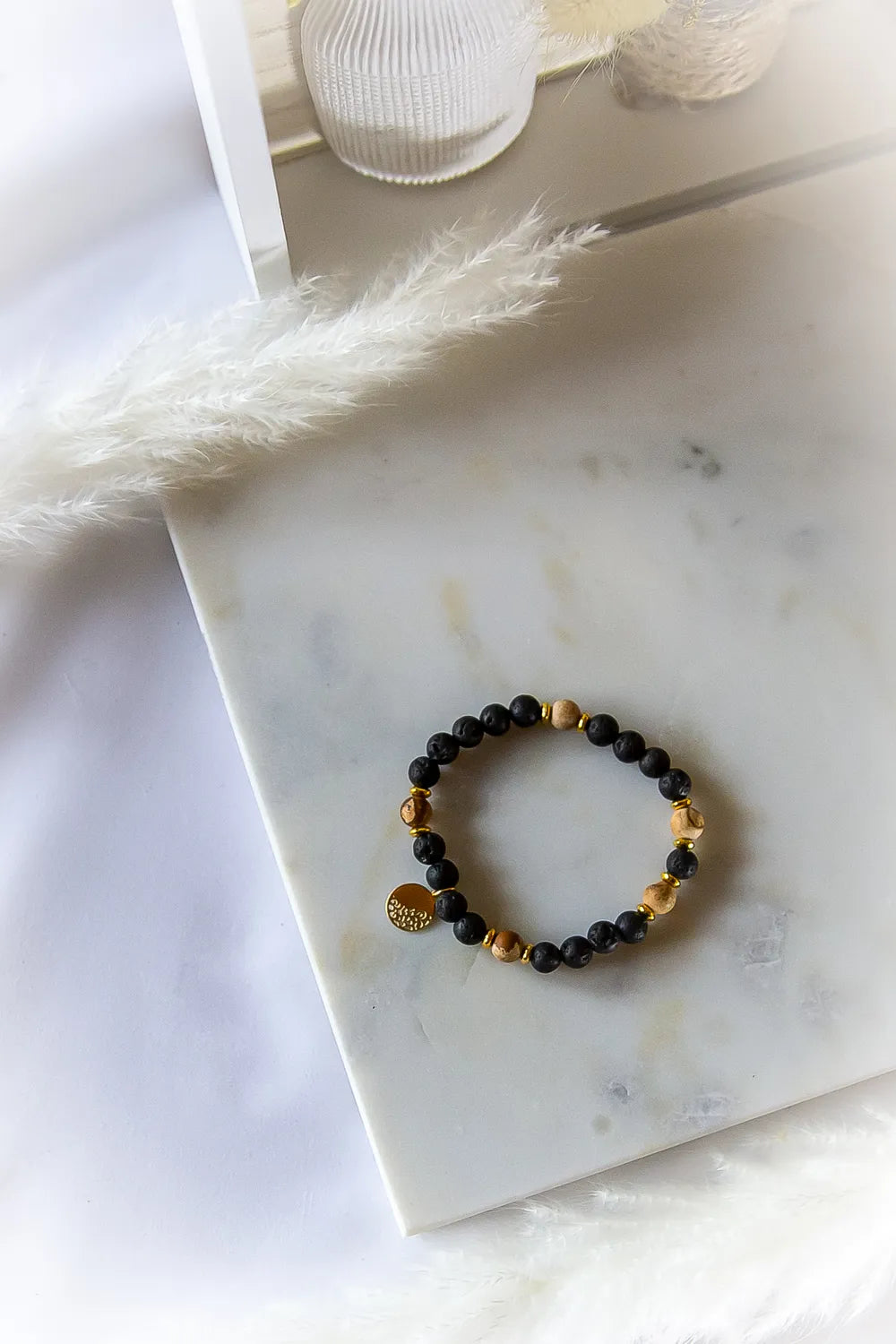 Ziemskie Ścieżki Bransoletka Jaspis Czarna Lawa z kamieni lawy i jaspisu na białym marmurze, ozdobiona złotym medalionem.