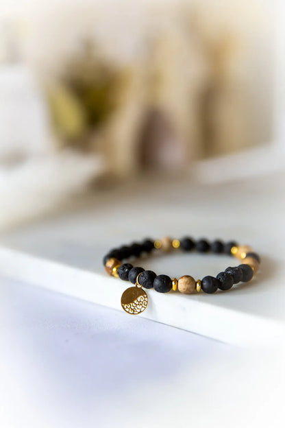 Ziemskie Ścieżki Bransoletka Z Jaspisu I Czarnej Lawy Złoto - zbliżenie na elastyczną bransoletkę z matowych, czarnych kamieni lawy i beżowych kamieni jaspisu, przeplatanych złotymi przekładkami. Złoty, okrągły medalik z ażurowym wzorem zwisa od przodu.
