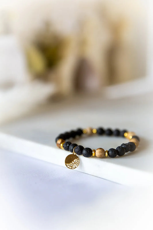 Ziemskie Ścieżki Bransoletka Z Jaspisu I Czarnej Lawy Złoto - zbliżenie na elastyczną bransoletkę z matowych, czarnych kamieni lawy i beżowych kamieni jaspisu, przeplatanych złotymi przekładkami. Złoty, okrągły medalik z ażurowym wzorem zwisa od przodu.