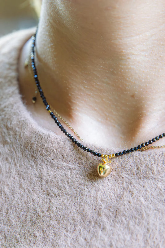 ZŁOTE JABŁKO - NASZYJNIK Z CZARNEGO TURMALINU. Naszyjnik z czarnymi koralikami turmalinu i złotym zawieszką w kształcie jabłka, na tle delikatnej, beżowej tkaniny.