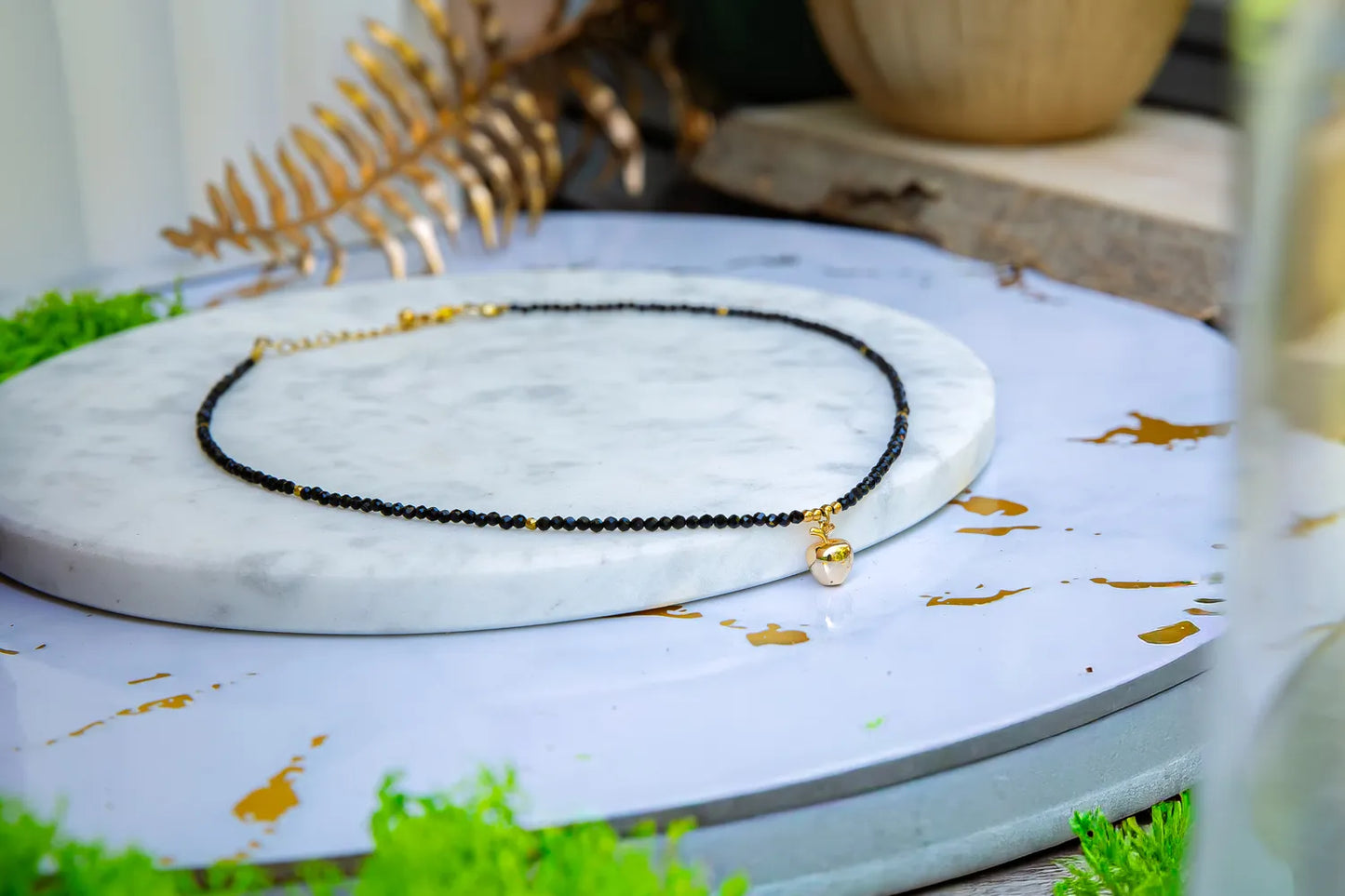 ZŁOTE JABŁKO - NASZYJNIK Z CZARNEGO TURMALINU, elegancki naszyjnik z czarnymi koralikami turmalinu i złotym zawieszką w kształcie jabłka, ułożony na marmurowym talerzu na tle zielonych liści i złotych elementów.