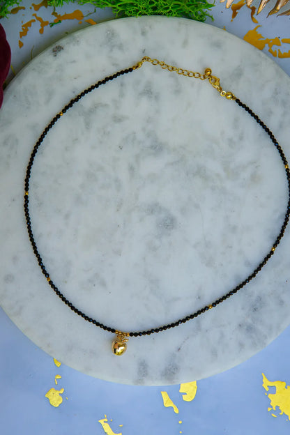 ZŁOTE JABŁKO - NASZYJNIK Z CZARNEGO TURMALINU, elegancki naszyjnik z czarnymi koralikami turmalinu i złotym jabłkiem, ułożony na marmurowym tle z dekoracjami.