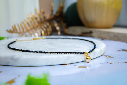 ZŁOTE JABŁKO - NASZYJNIK Z CZARNEGO TURMALINU, elegancki naszyjnik z czarnymi koralikami turmalinu i złotym jabłkiem, ułożony na marmurowym podstawce z dekoracją w tle.