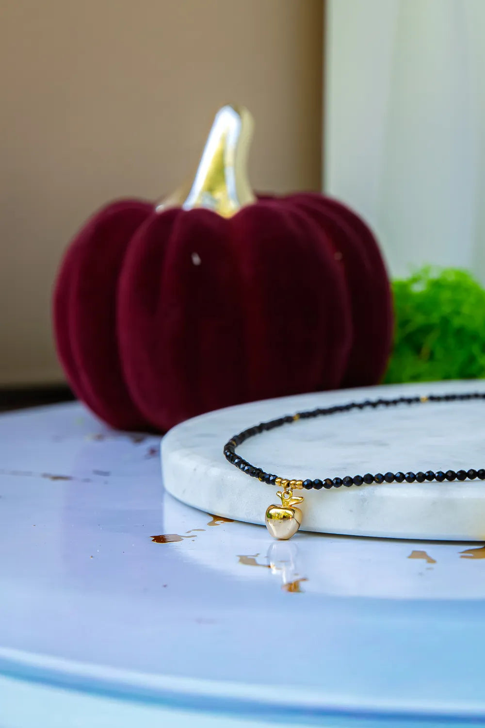 ZŁOTE JABŁKO - NASZYJNIK Z CZARNEGO TURMALINU, elegancki naszyjnik z czarnymi koralikami turmalinu i złotym zawieszką w kształcie jabłka, ułożony na marmurowym talerzu z czerwoną dynią w tle.