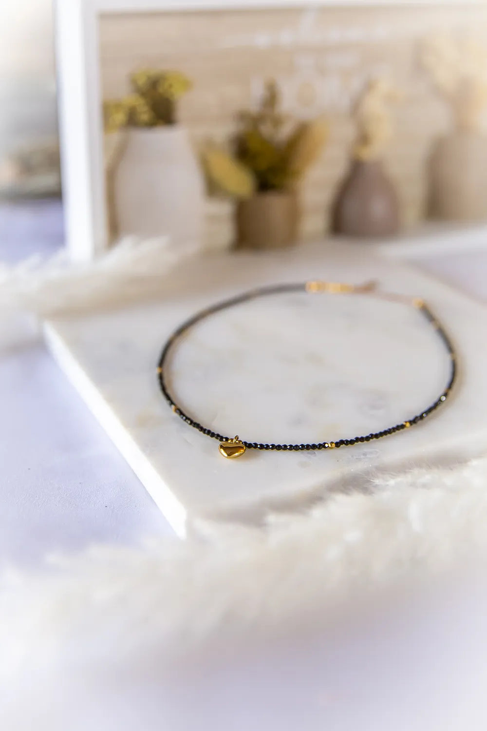 Złote Serduszko Naszyjnik Turmalin: delikatny naszyjnik z czarnych turmalinów i złotych kuleczek, z zawieszką w kształcie złotego serduszka, na tle bluru z ozdobnymi wazonami.