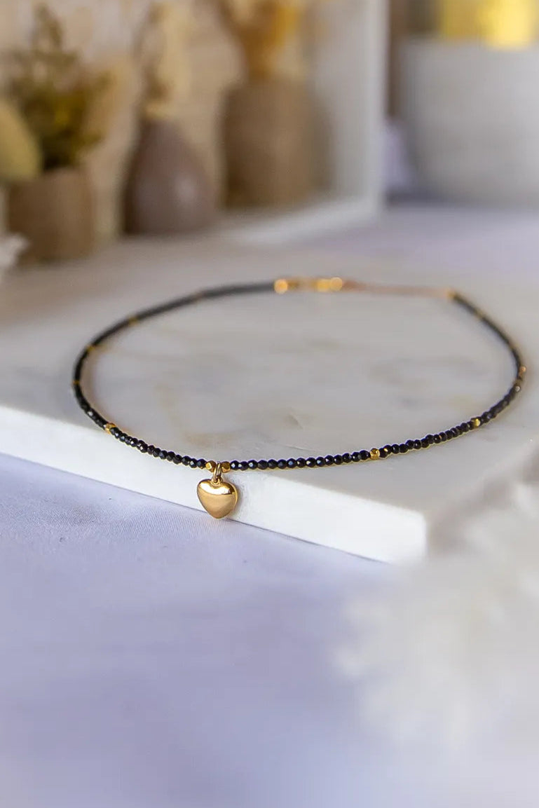 Złote Serduszko Naszyjnik Turmalin z drobnymi czarnymi kamieniami i złotymi akcentami, wyeksponowany na marmurowej płycie. Dyskretne tło z ozdobnymi wazonami dodaje elegancji.