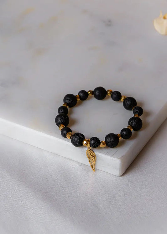 Złote Skrzydło Bransoletka Czarna Lawa Hematyt z złotym skrzydełkiem, na białej marmurowej płytce.