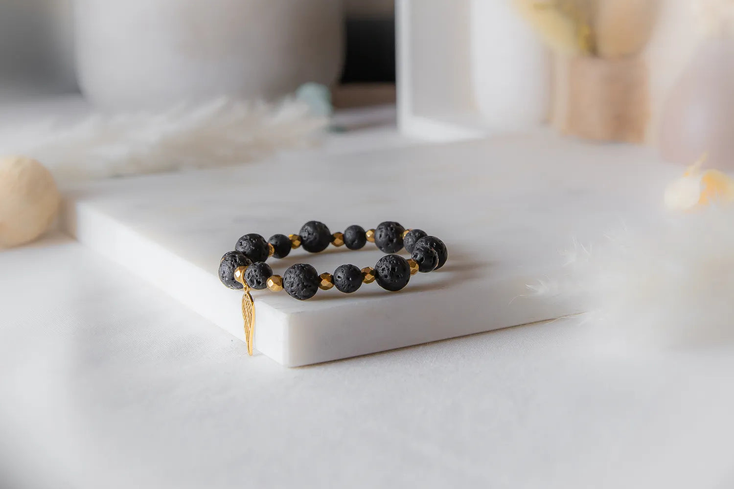 Złote Skrzydlo Bransoletka Czarne Kamienie z złotą zawieszką skrzydła, na białej marmurowej powierzchni, delikatne tło z piórami.