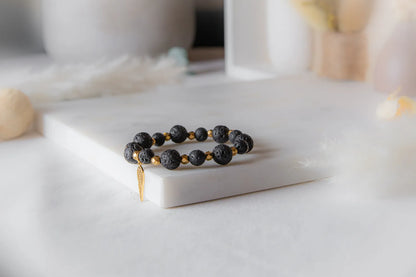 Złote Skrzydlo Bransoletka Czarne Kamienie z złotą zawieszką skrzydła, na białej marmurowej powierzchni, delikatne tło z piórami.