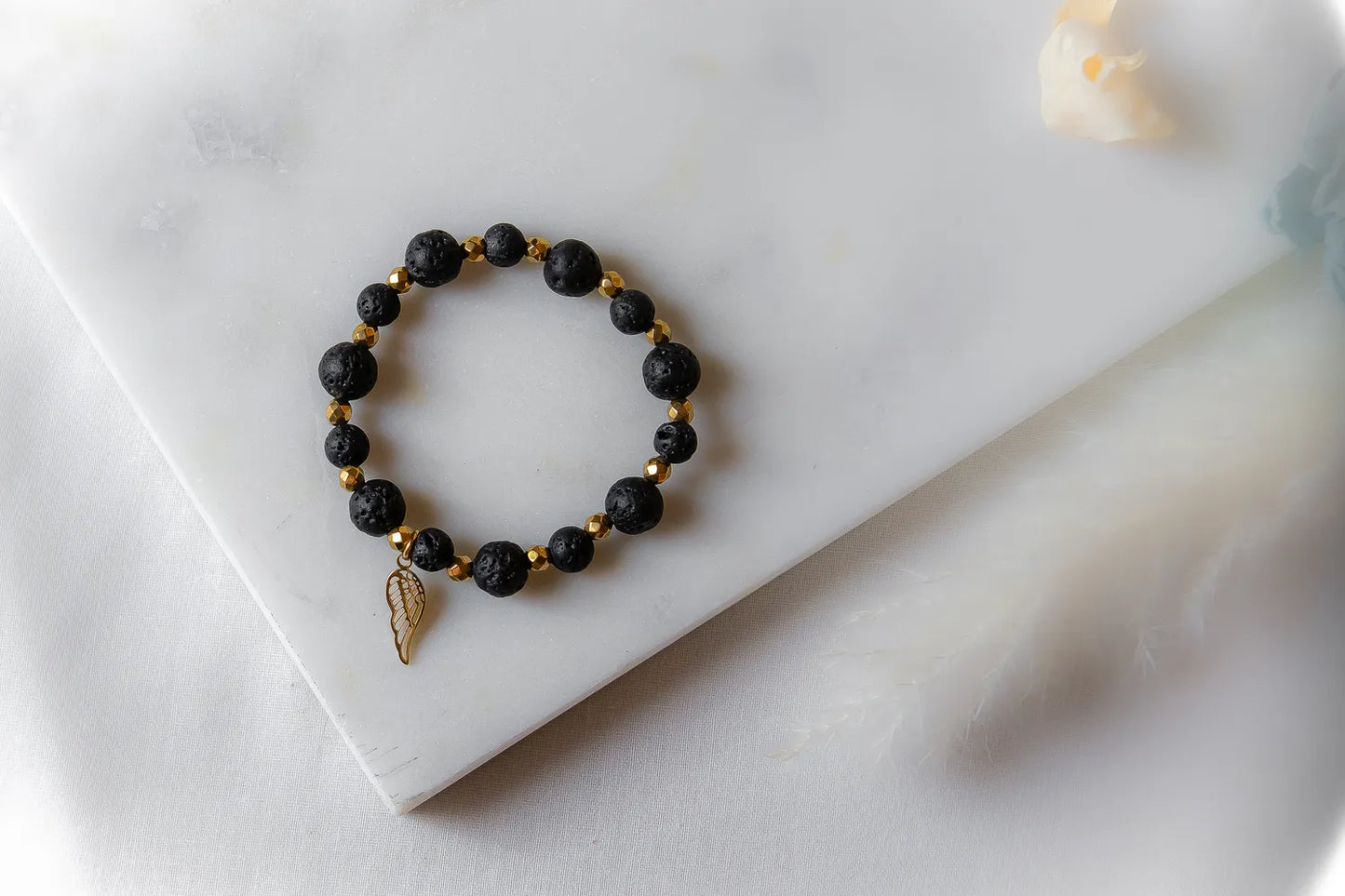 Złote Skrzydło Bransoletka Czarne Kamienie: czarne, fasetowane kamienie lawy i złote przekładki, z zawieszką skrzydło, na białym tle.