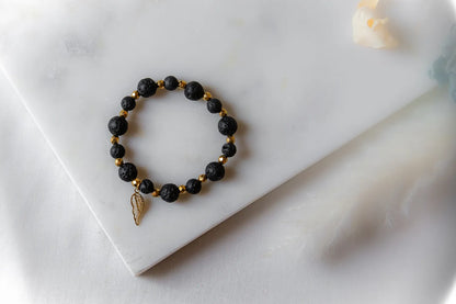 Złote Skrzydło Bransoletka Czarne Kamienie: czarne, fasetowane kamienie lawy i złote przekładki, z zawieszką skrzydło, na białym tle.