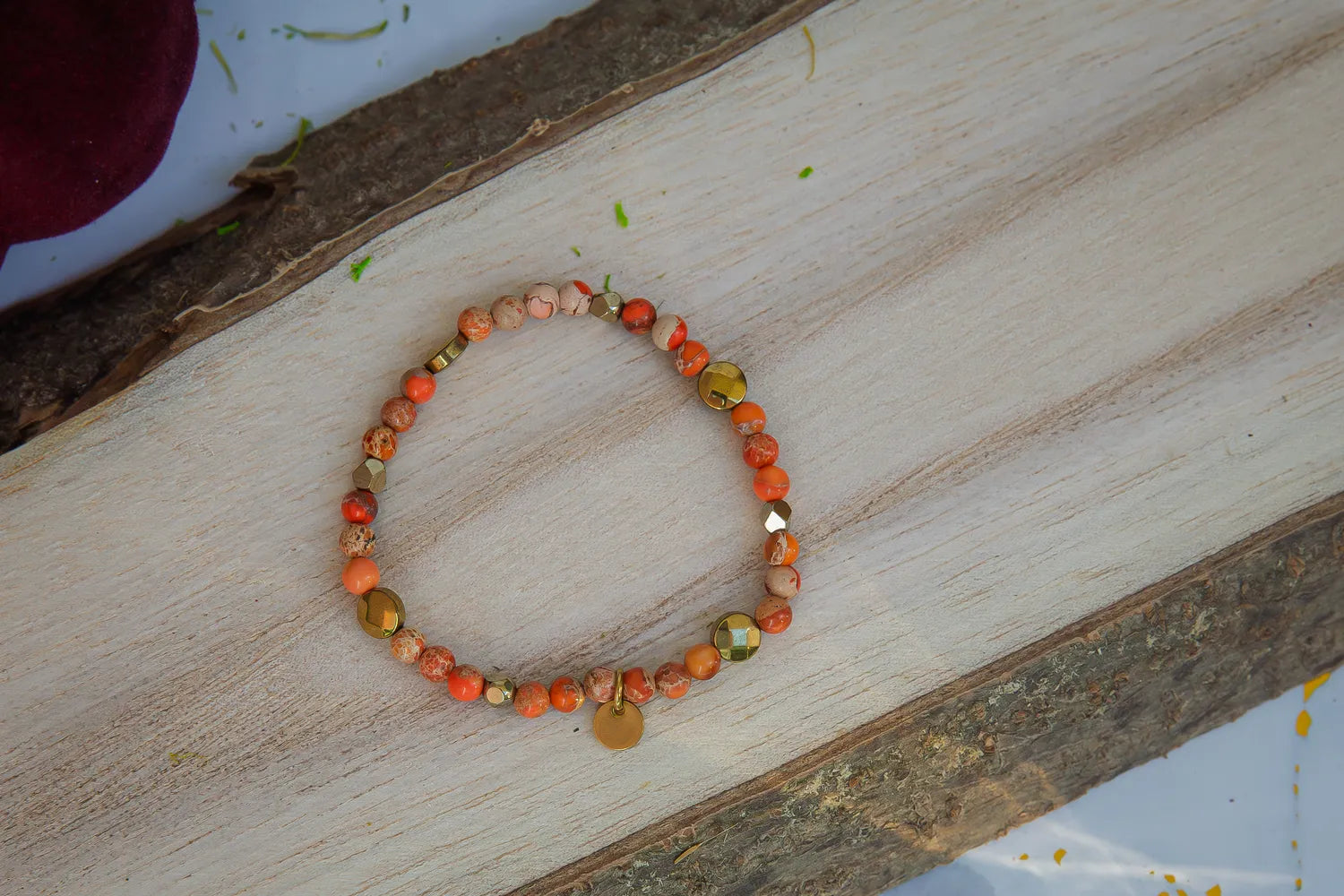 ZŁOTO PAŹDZIERNIKA - BRANSOLETKA Z JASPISU CESARSKIEGO I HEMATYTU, prezentująca się na drewnianym tle. Biżuteria składa się z kolorowych kamieni jaspisu cesarskiego i hematytu, ułożonych w elegancki wzór.