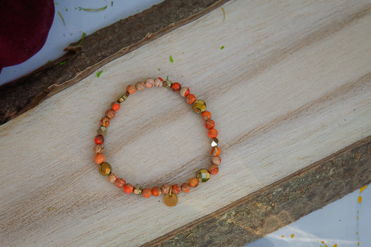 ZŁOTO PAŹDZIERNIKA - BRANSOLETKA Z JASPISU CESARSKIEGO I HEMATYTU, prezentująca się na drewnianym tle. Biżuteria składa się z kolorowych kamieni jaspisu cesarskiego i hematytu, ułożonych w elegancki wzór.