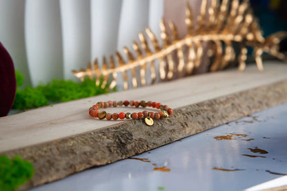 ZŁOTO PAŹDZIERNIKA - BRANSOLETKA Z JASPISU CESARSKIEGO I HEMATYTU, prezentująca się na drewnianym tle z zielonymi dodatkami. Biżuteria składa się z kolorowych kamieni i małych złotych elementów.