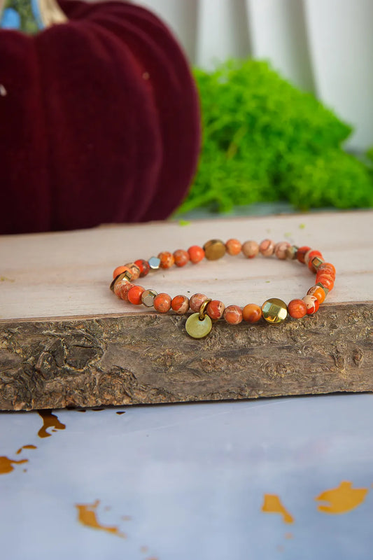 ZŁOTO PAŹDZIERNIKA - BRANSOLETKA Z JASPISU CESARSKIEGO I HEMATYTU, prezentująca się na drewnianym tle z ciepłymi, pomarańczowymi koralikami jaspisu cesarskiego i ciemnymi koralikami hematytu, z dodatkowym elementem w kształcie liścia.